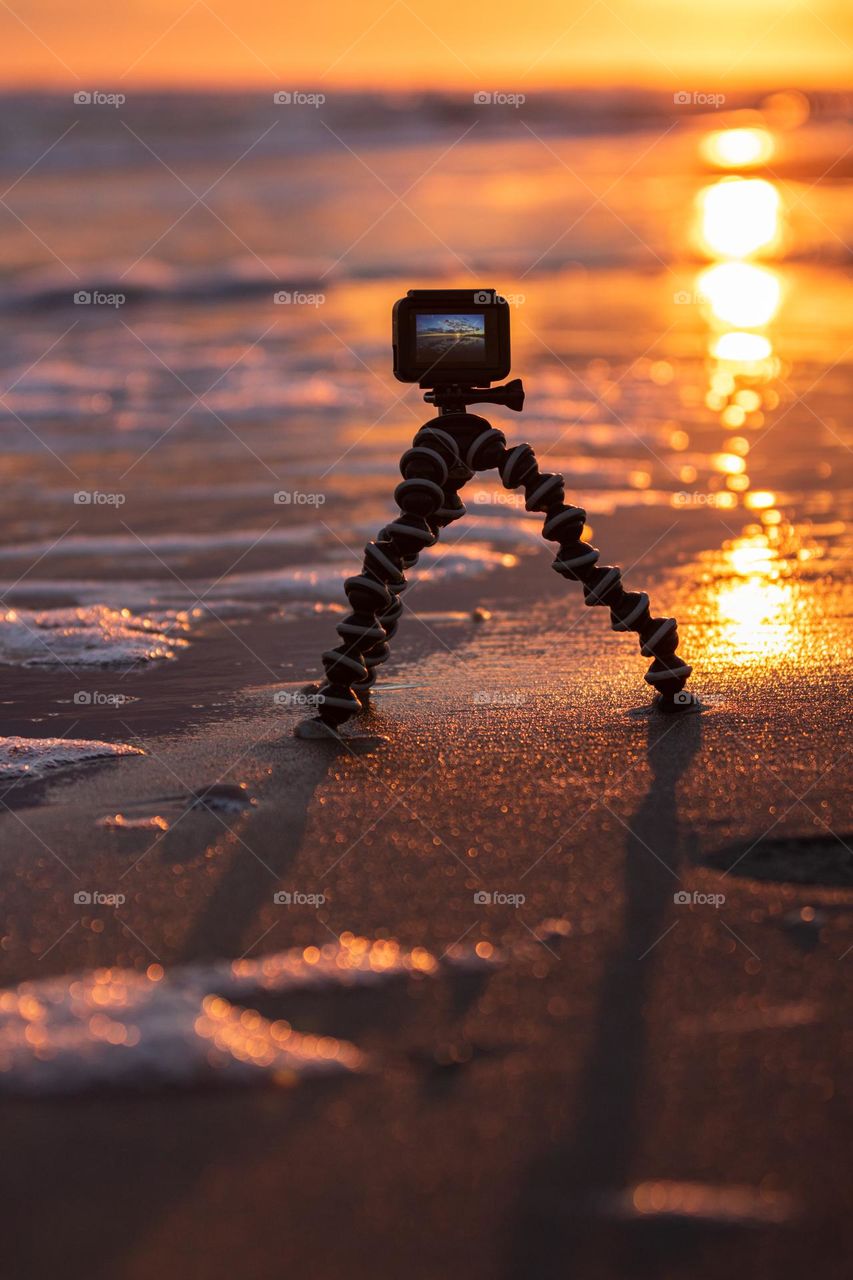 A GoPro action camera set up on a mini tripod capturing a beautiful sunset on a beach.