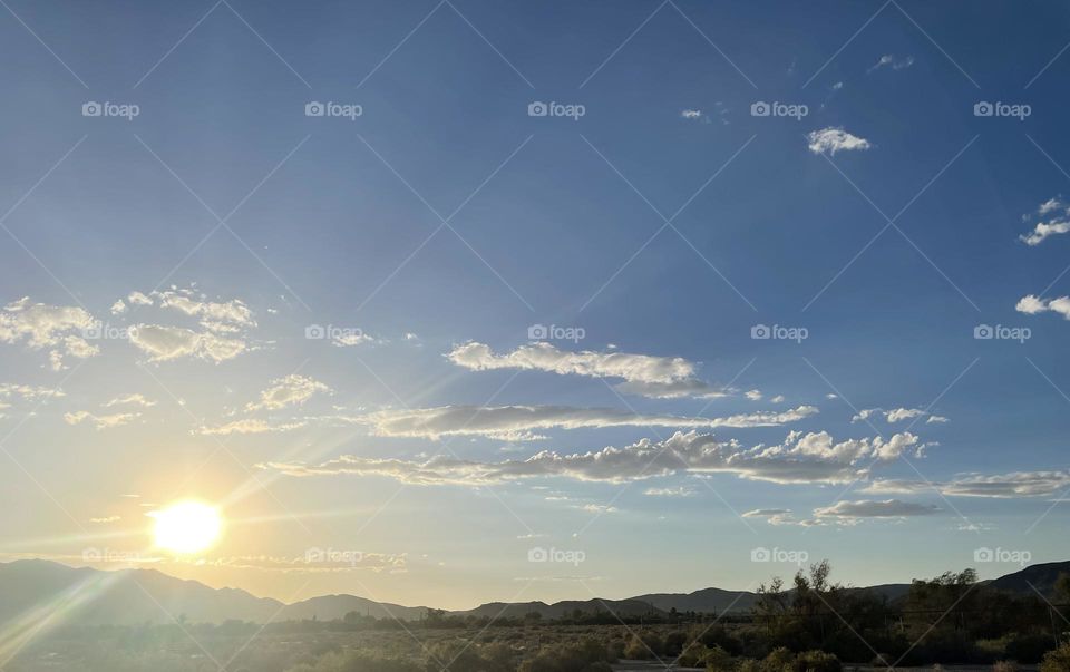 A blue sky with the sun shining and mountains with clouds above it all. 