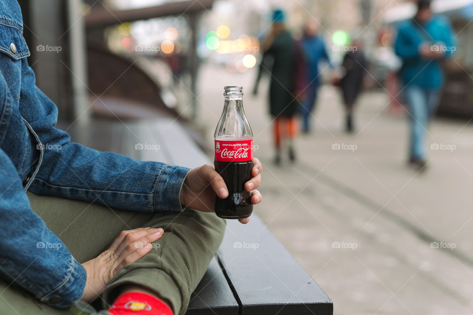 a guy sits on the street, holds a bottle of Coca-Cola in his hands, opens it.  the guy is dressed in denim clothes.