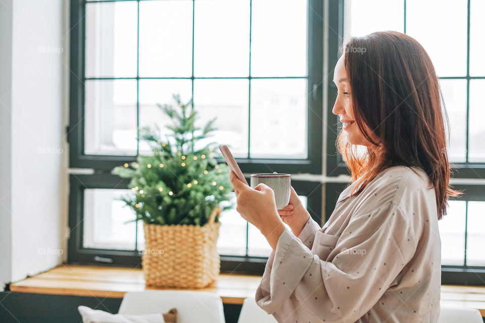 Adult young woman in pajamas with coffee mug in hand using mobile phone in room with Christmas tree at home 