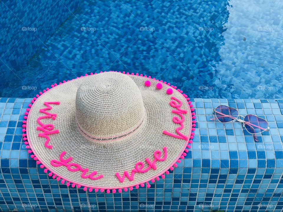 Summer hat with the words “wish you were here” and sunglasses on the edge of a pool with turquoise water