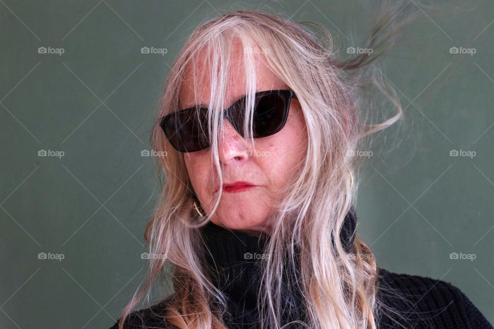 A senior woman with long blonde tousled hair, sunglasses and gold earrings against a green background