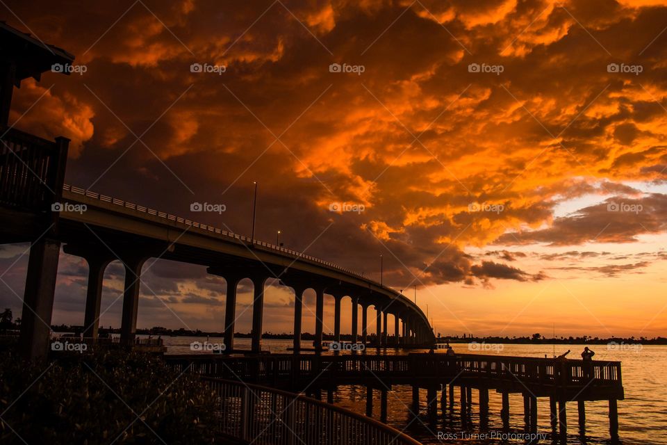The bridge and fishing pier appear as silhouettes against the sunset sky. The orange sky is reflected in the clouds directly overhead.