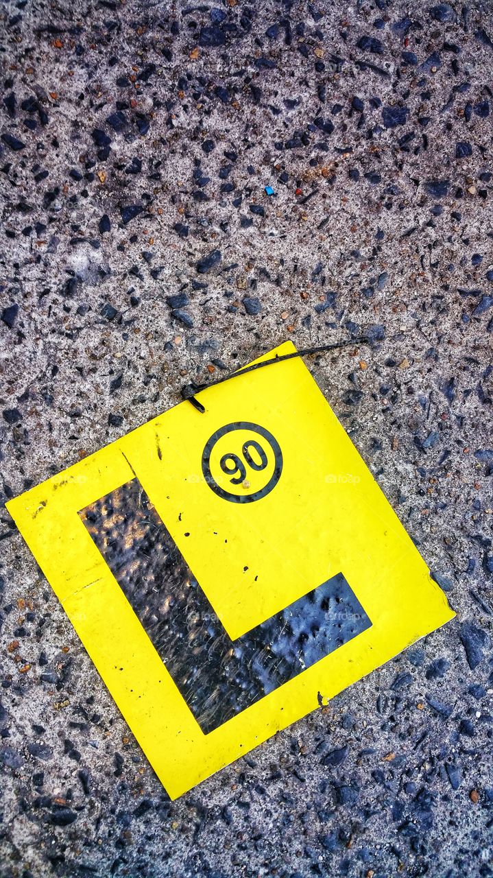 A bright yellow vehicle L-plate badge that has fallen onto a highly textured concrete floor. The circled '90' insignia denotes a maximum travel speed limit of 90 km/hr.