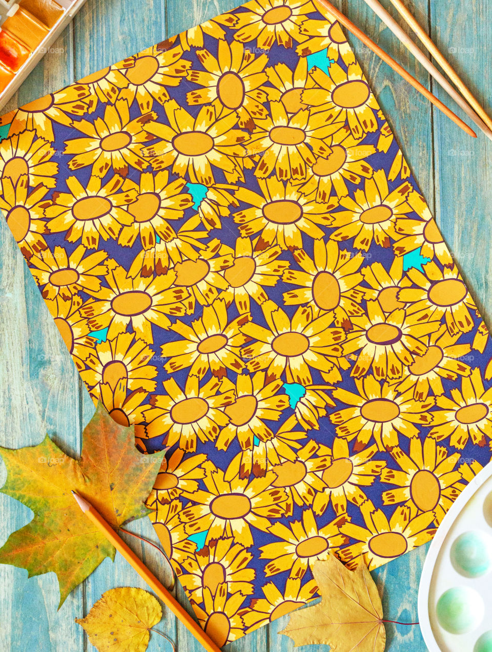 Creative work, paint drawing. A table with a decorative repeating pattern with daisies, next to which are brushes, watercolor and gouache paints, a palette and dry autumn colored leaves.