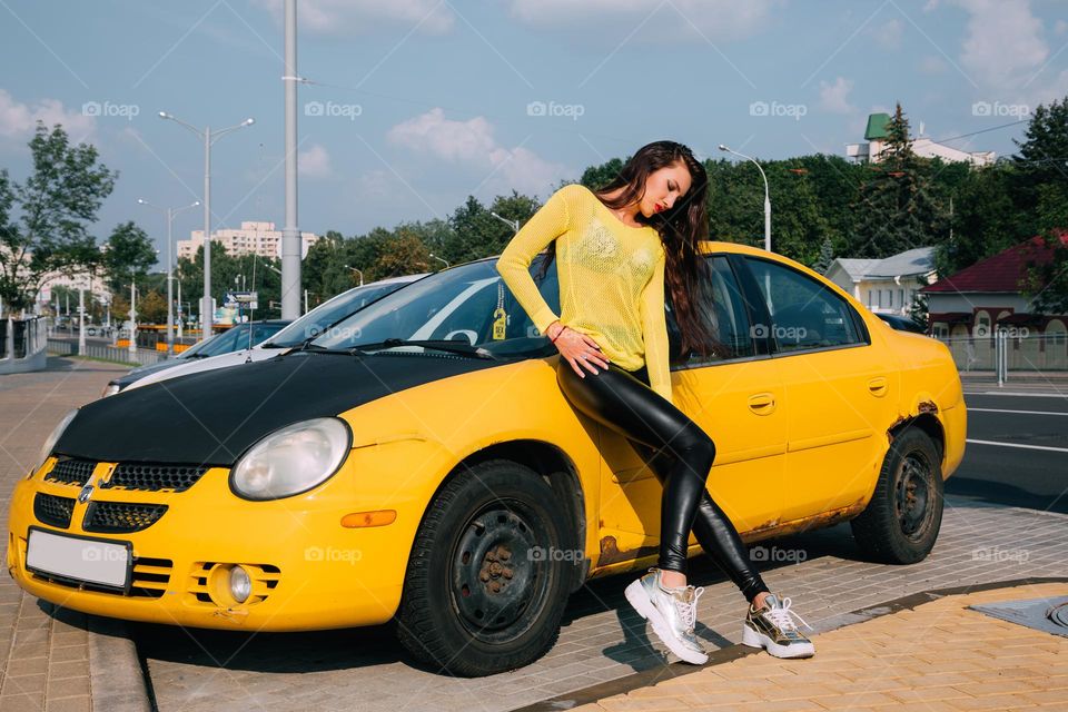 Pretty brunette, wearing a yellow sweater, shiny black leggings and golden sneakers, leans against yellow car