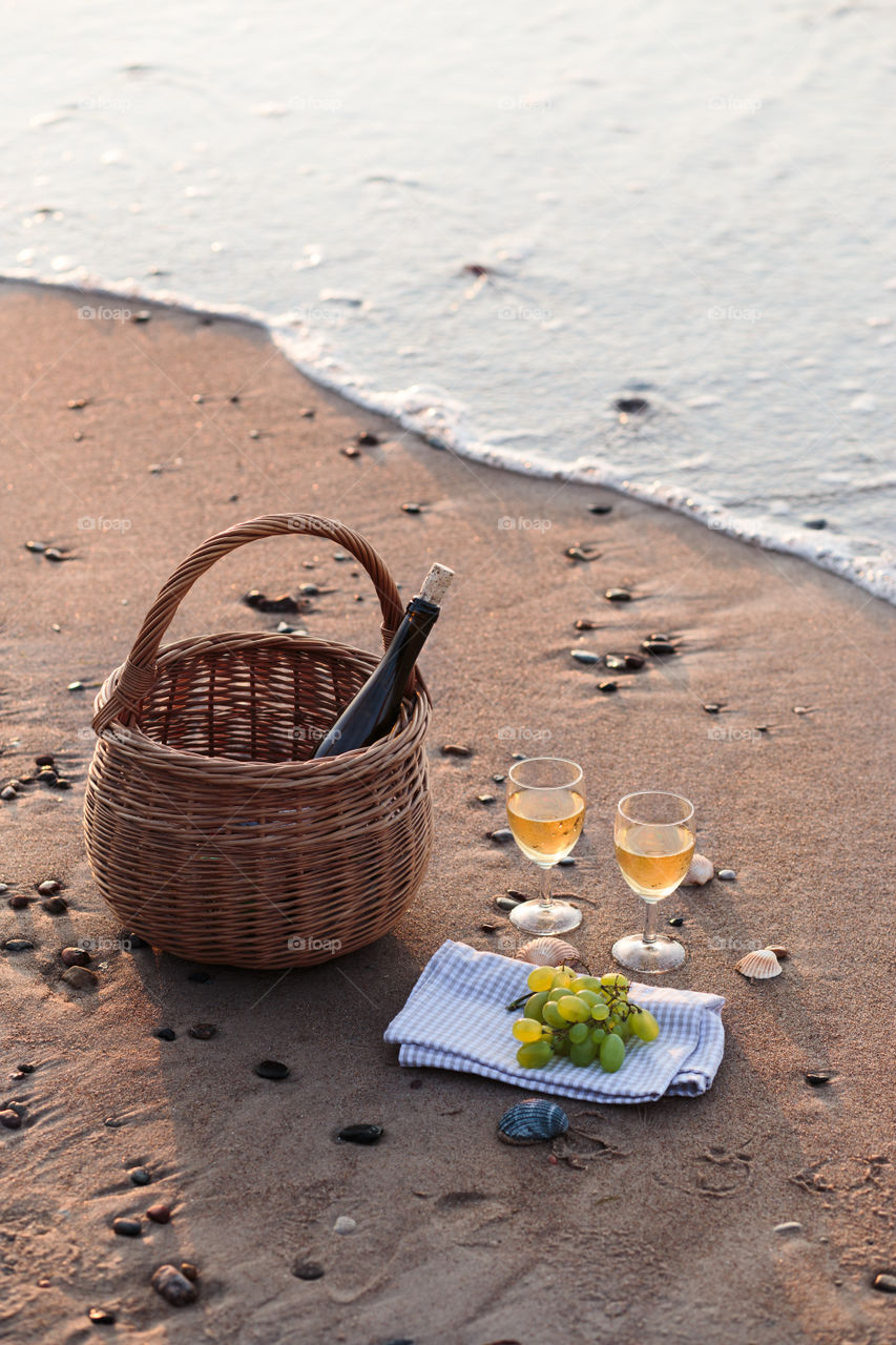 Two wine glasses with white wine standing on sand, on beach, beside grapes and wicker basket with bottle of wine. Sea waves in the background