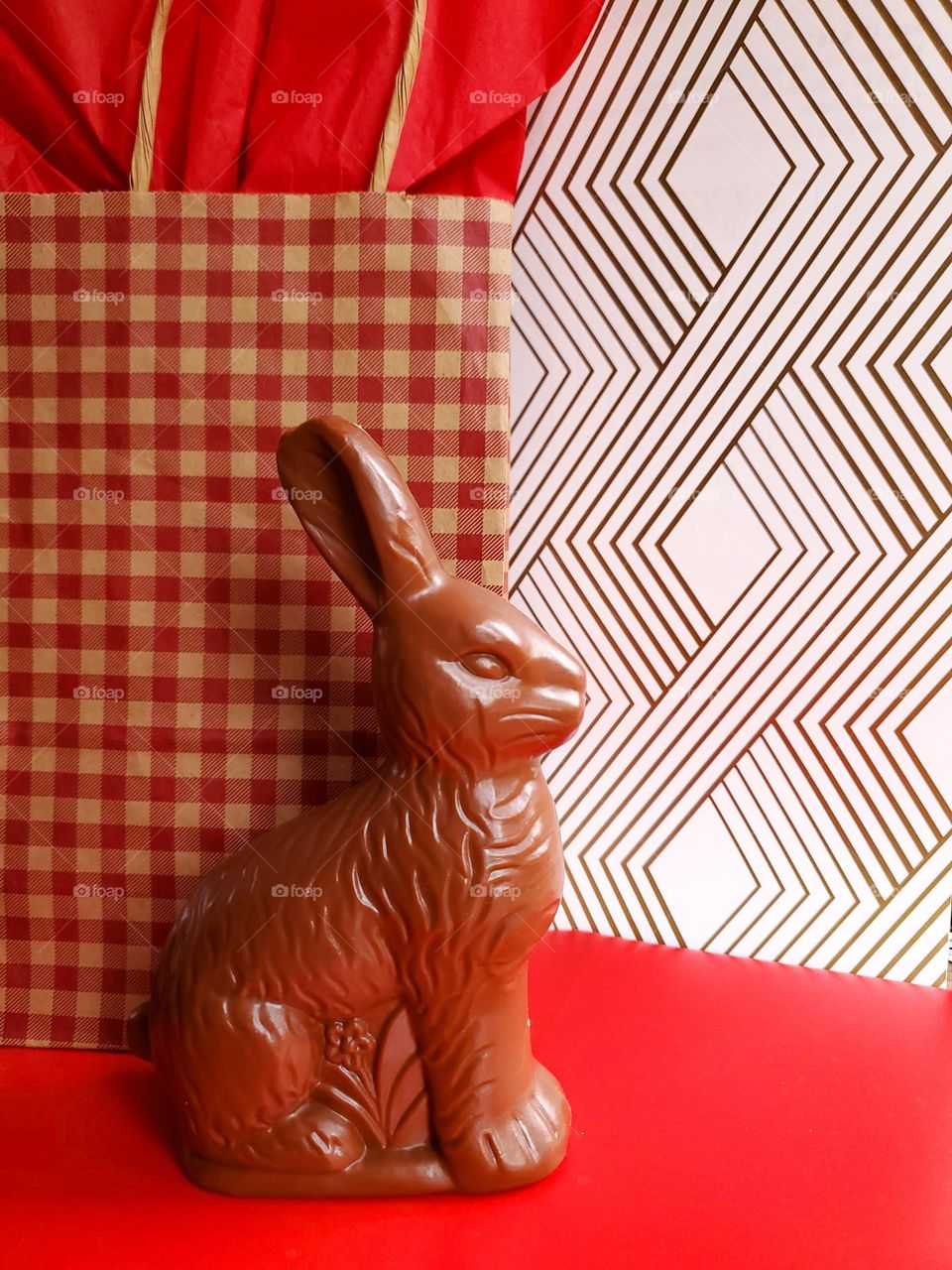 Faux chocolate bunny in front of a red and light brown plad gift bag with a gold and white multiple diamond shape background and a red surface.