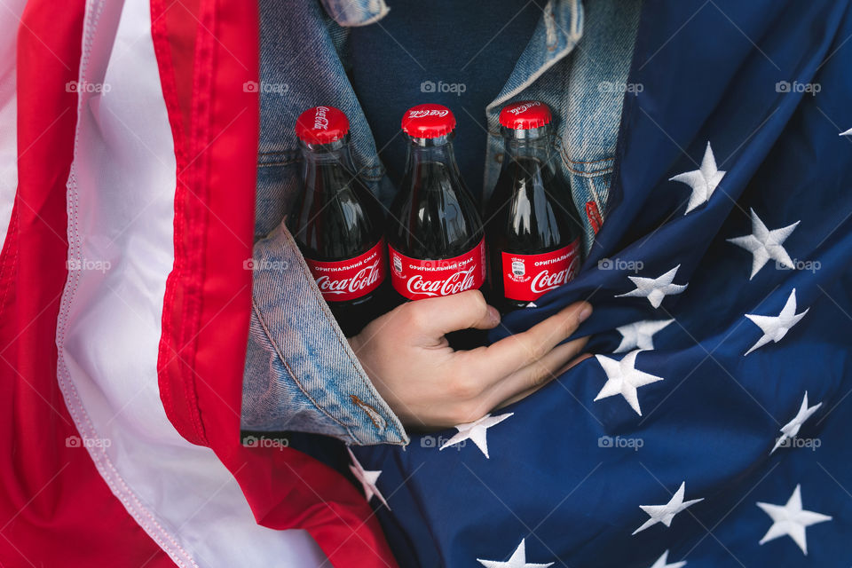 a teenage guy sitting on a bench holding a Coca-Cola in his hands.  the guy is shrouded in the American flag