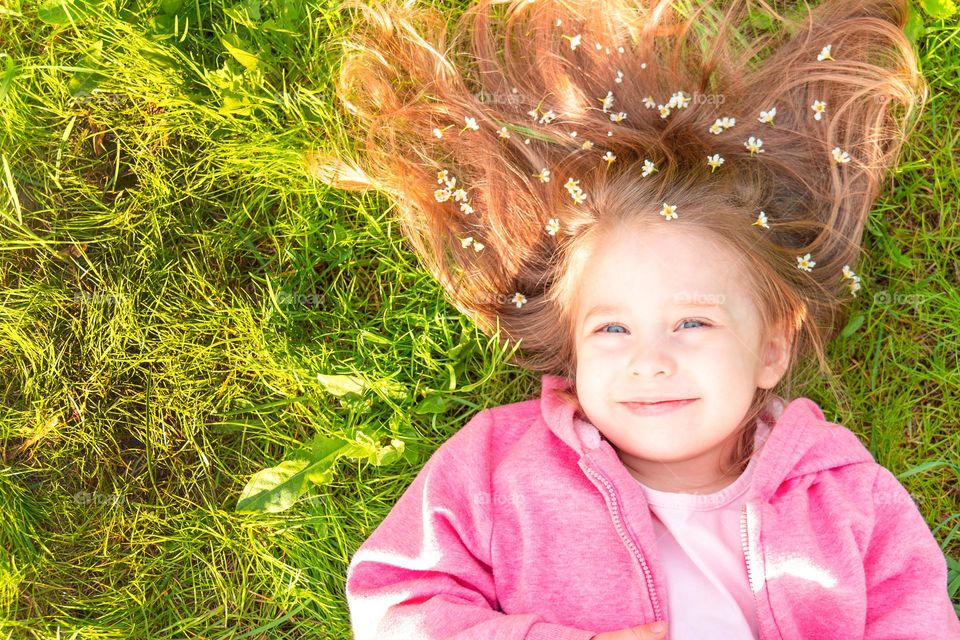 Top view of smiling cute girl with flowers in hair lying on green grass and looking at camera