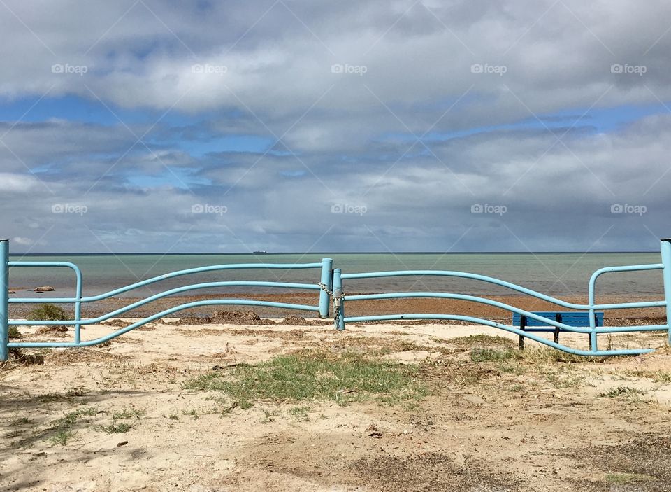 Metal wavy metal gate in aqua marine blue padlocked barring entry of vehicles onto tropical beach with ocean and cloudy sky in the horizon 