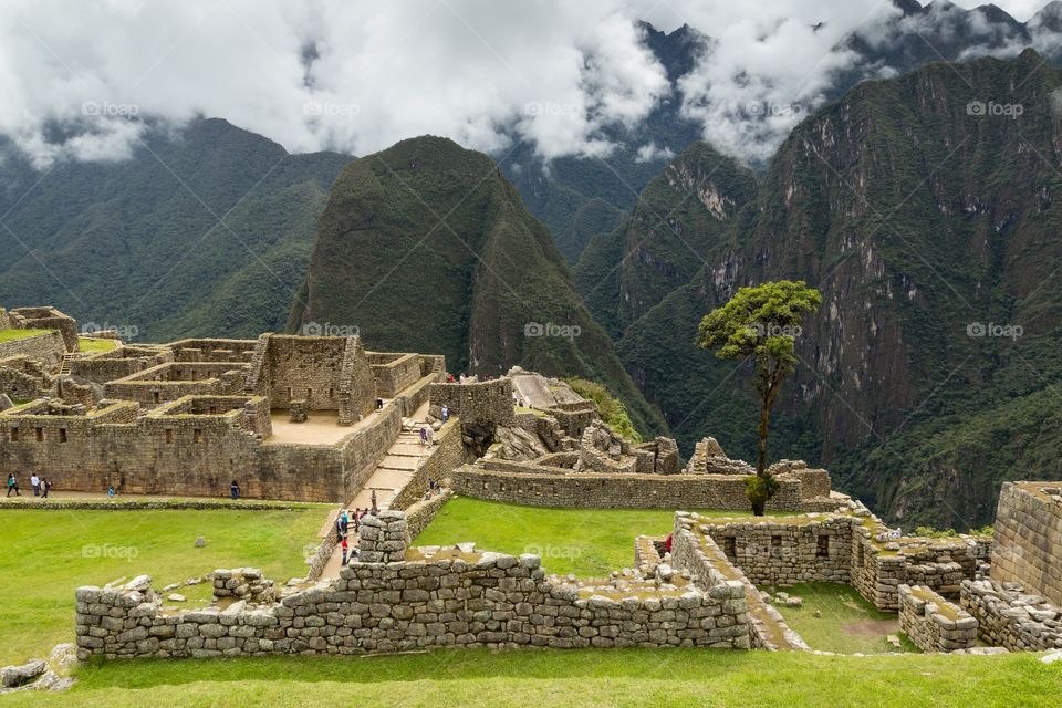 View towards Machu Picchu ruin. Machu Picchu ruins in foreground, steep mountains in the background. Stone structures. Cloudy sky