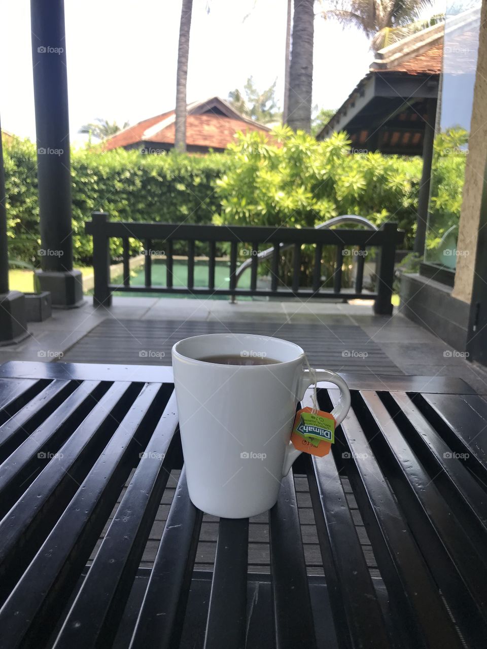 Hot tea cup were left on the black table on the porch,looking directly out there was a black wooden railing,a swimming pool with blue water.some coconut trees planted around,fence made of green trees,veranda floor is lined with black stone