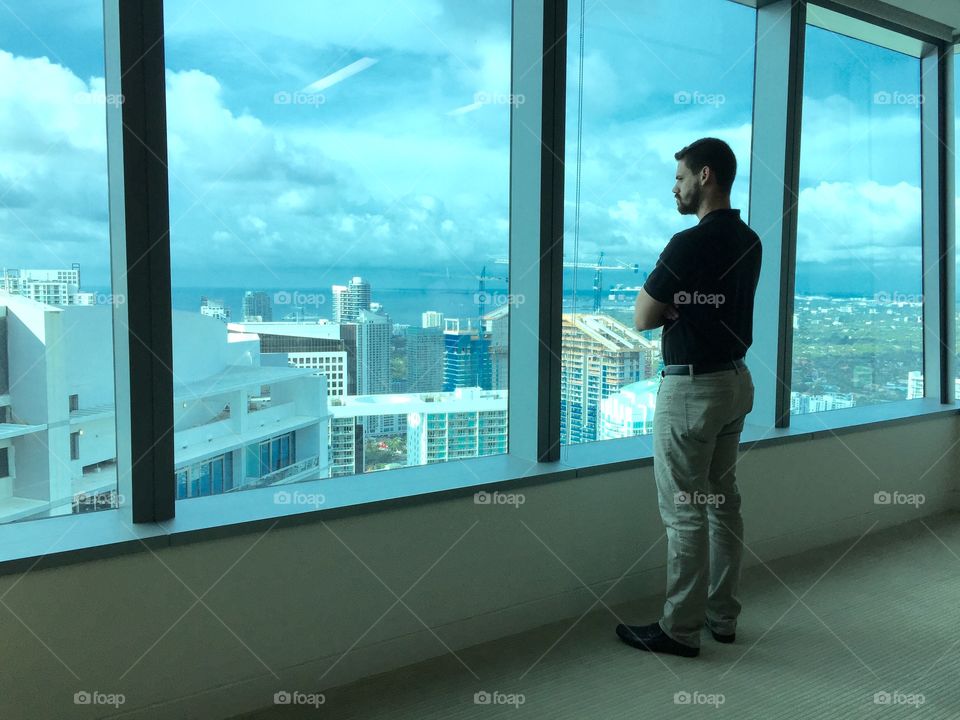 Contemplation . Young man standing by panoramic window in office looking outside
