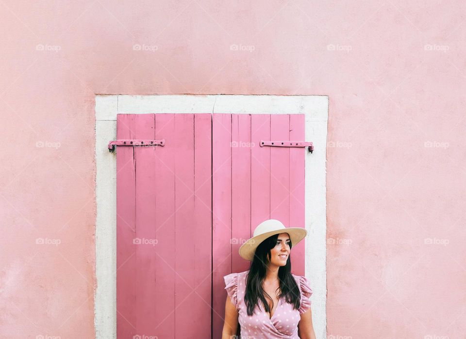 Portrait of young woman wearing stylish summer clothes and sun hat, standing in front of window with pink shutters