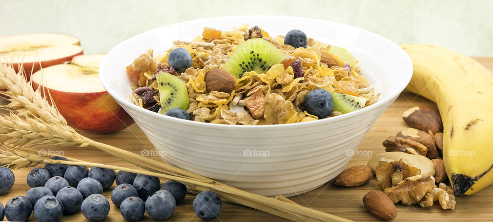 A bowl of wholegrain  cereal topped with blueberries and kiwifruit pieces and surrounded by banana, apples, blueberries, nuts and wheat stalks.