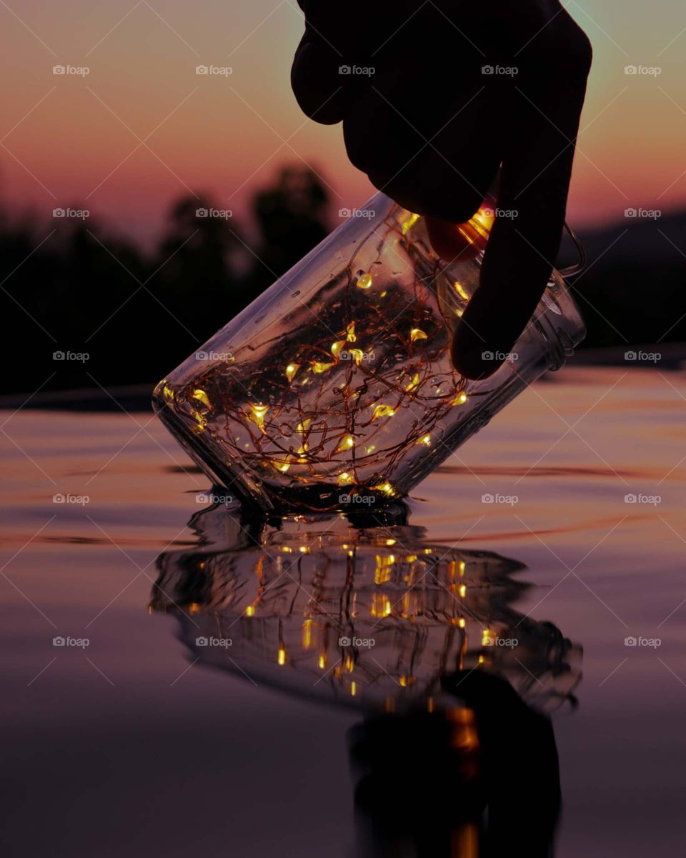 person holding string lights on clear glass jar above body of water.