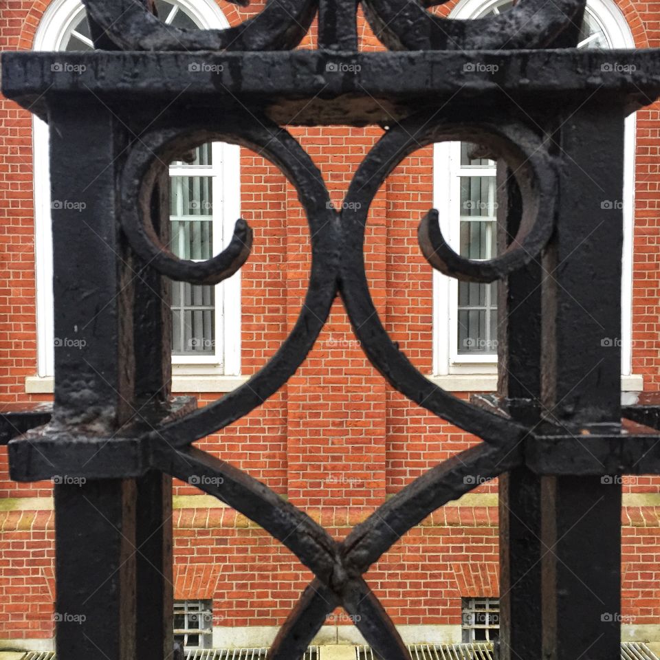 Decorative wrought iron work, painted black, on a fence located outside of the post office in Salem, Massachusetts. The background is a brick building with windows accented with white paint.