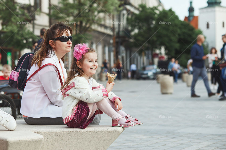 Mother and daughter spending time together, sitting in the center of town, little smiling girl holding and eating ice cream. Candid people, real moments, authentic situations