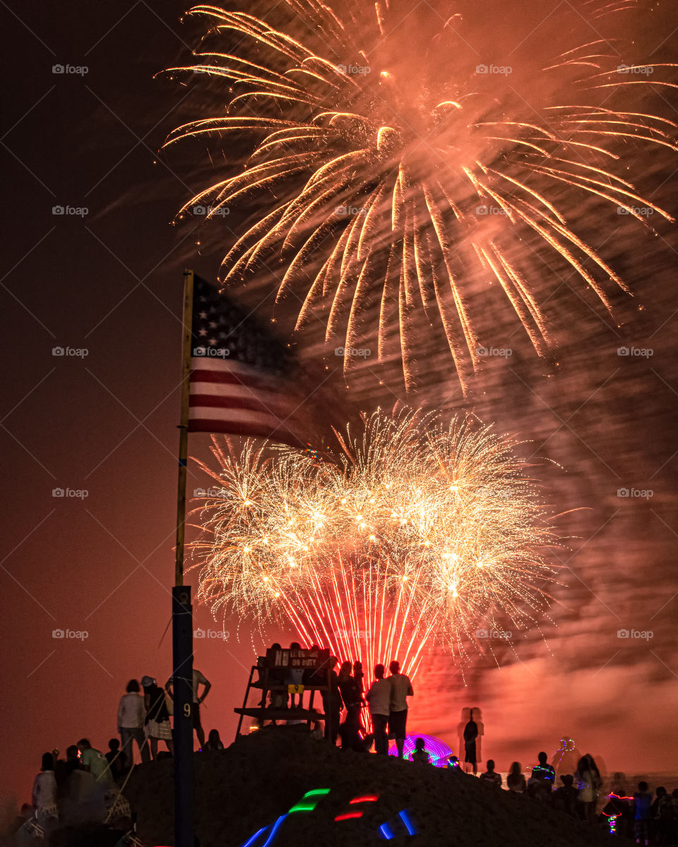 An American flag waving in the wind, as a brilliant fireworks display lights up the sky.