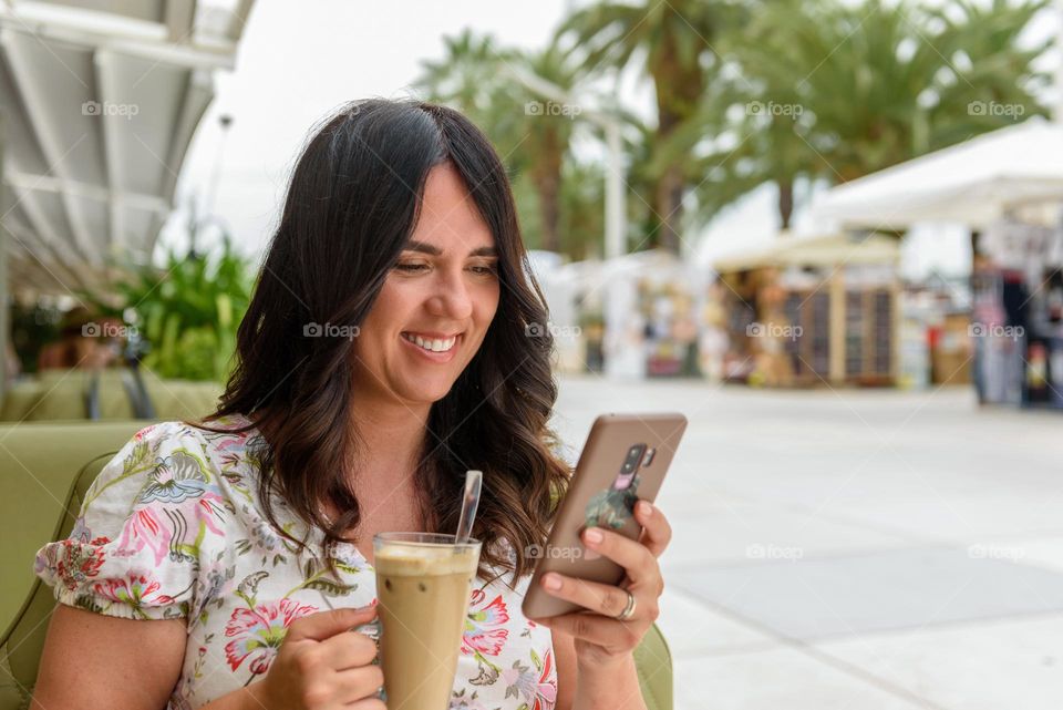 Happy smiling young woman holding mobile phone and texting while sitting outdoor in bar in city