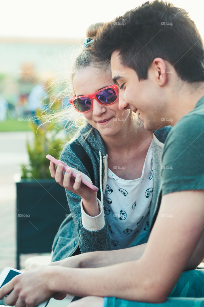 Couple of friends, teenage girl and boy, having fun using smartphones sitting in center of town, spending time together