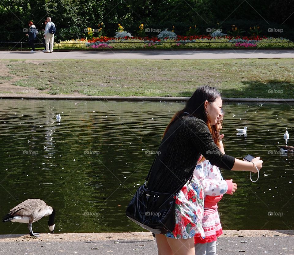 A mother and her daughter take a selfie while visiting an urban park with waster in the background on a sunny summer day. 