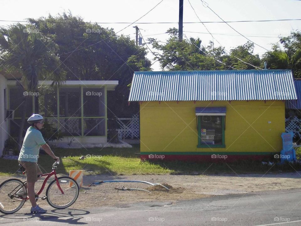 A young man wearing a green and white striped polo and helmet rides a bicycle on a paved road. The yellow building with a tin roof stand out on this leisurely ride. 