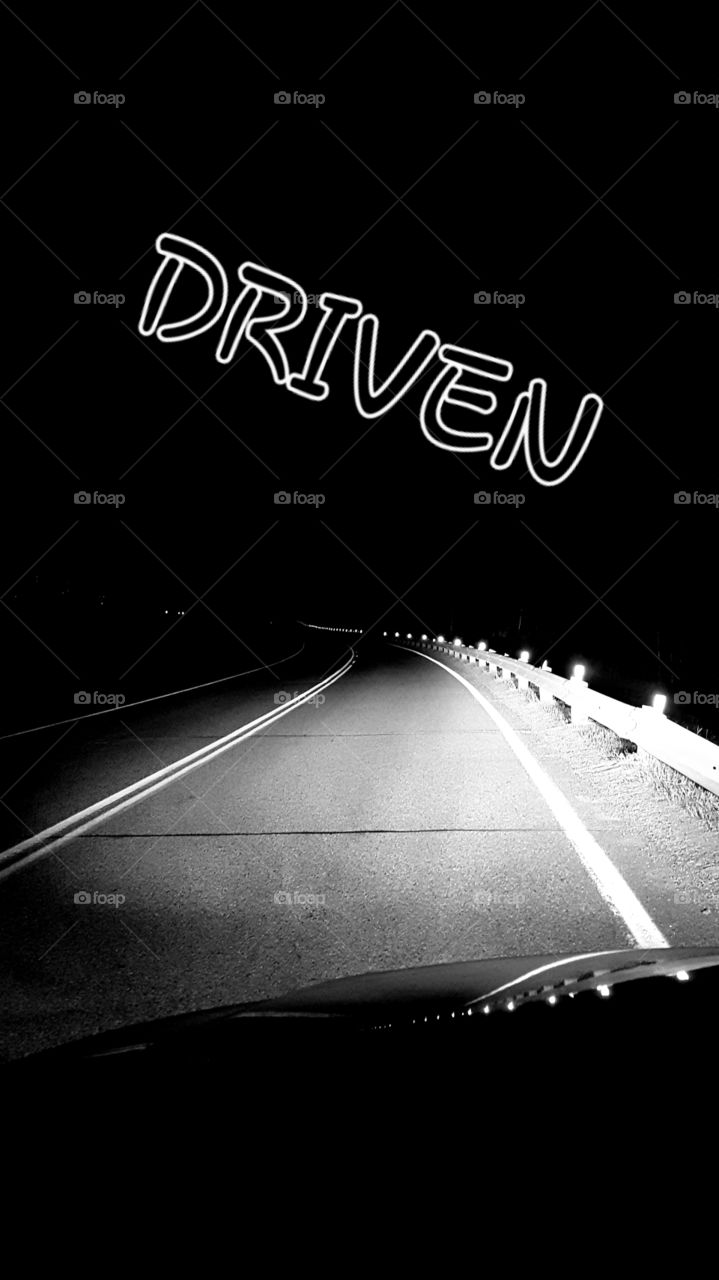 Black & White Vignette of a vehicle lights on a paved highway and guard rails at night with the word "driven".