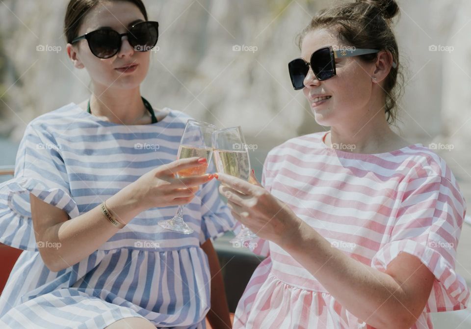 Two beautiful young and happy brunettes in sunglasses clink glasses of champagne in a boat in the sea against the backdrop of blurred rocks on a sunny summer day, close-up side view.