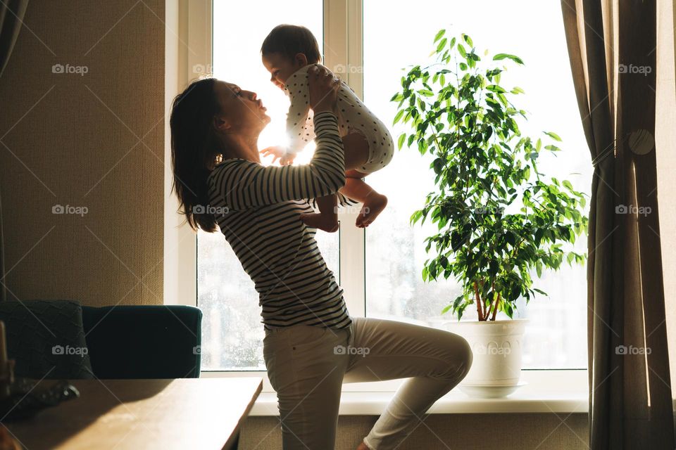 A young mother woman holding her little daughter baby girl in her arms by the window in the kitchen at home in the background light