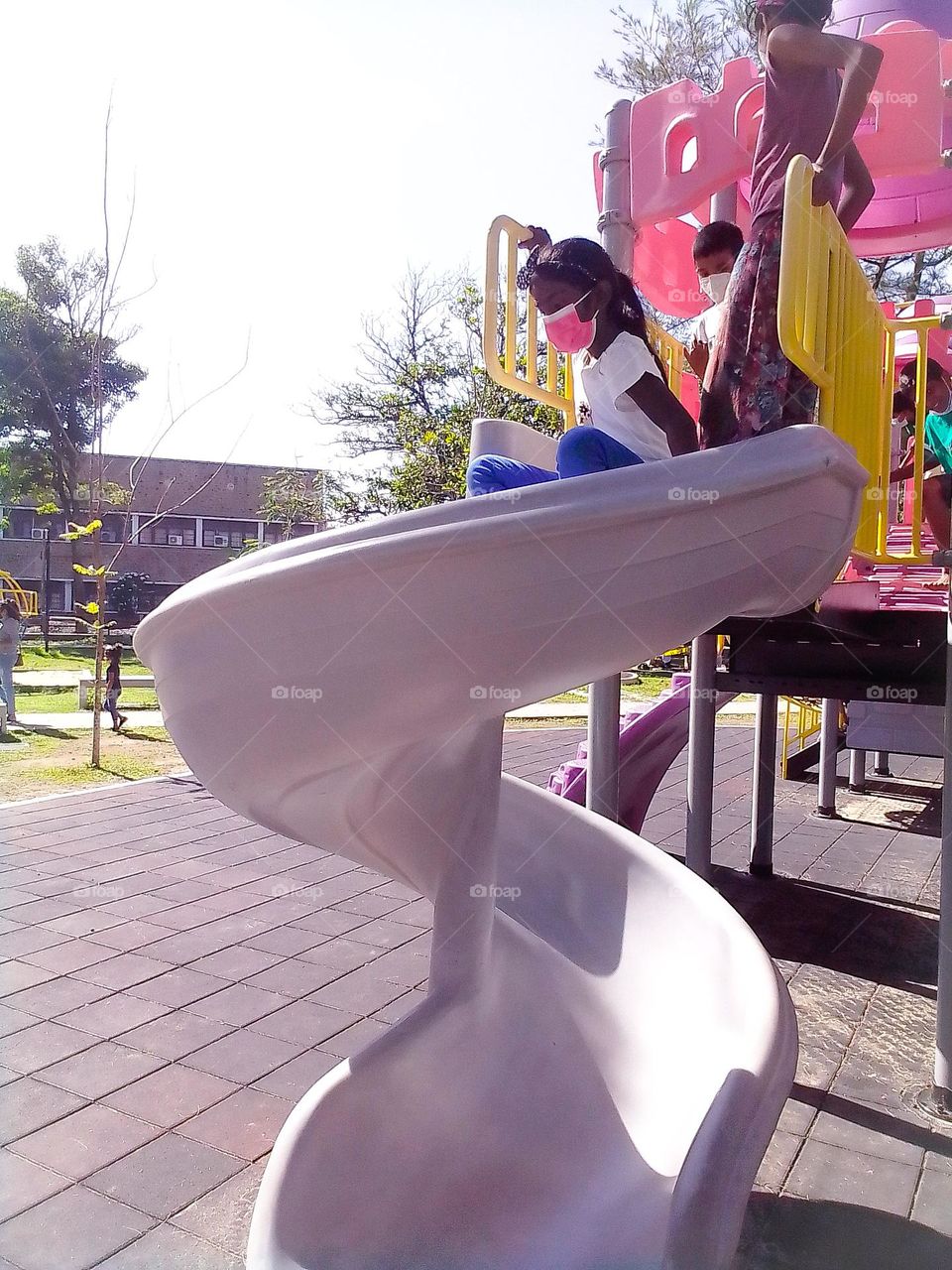 This is a capture of a children's park and children are playing. There is a white colour curved board and one girl is coming through it. She is wearing a pink colour mask.