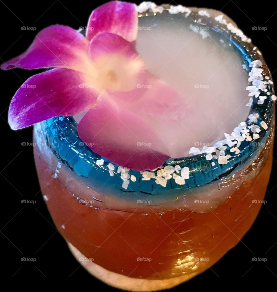 A vibrant pink rose cocktail in an aqua blue rimmed glass sprinkled with coarse white salt flakes, topped with a delicate orchid blossom and swirling with a dreamy mist of fog.