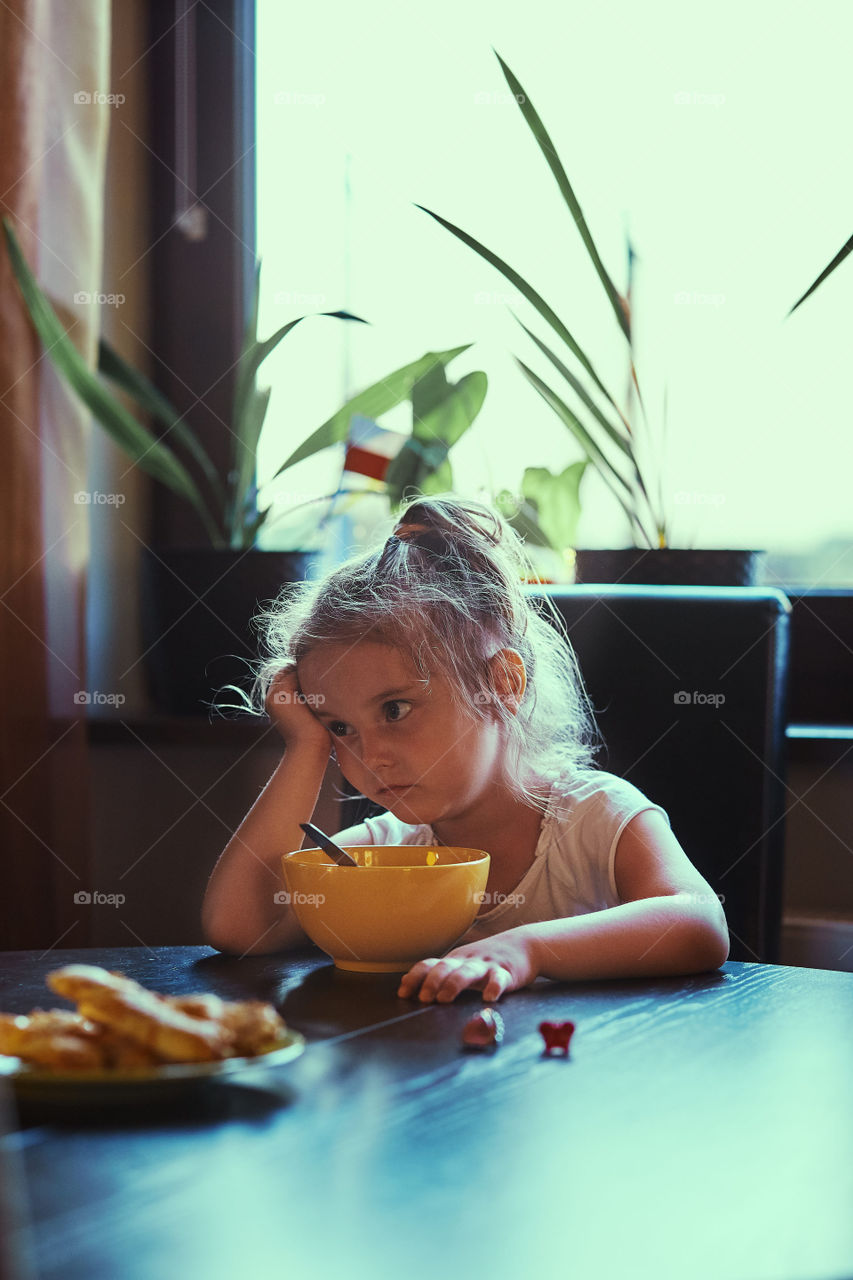 Little bored cute girl eating a breakfast, sitting by the table. Candid people, real moments, authentic situations