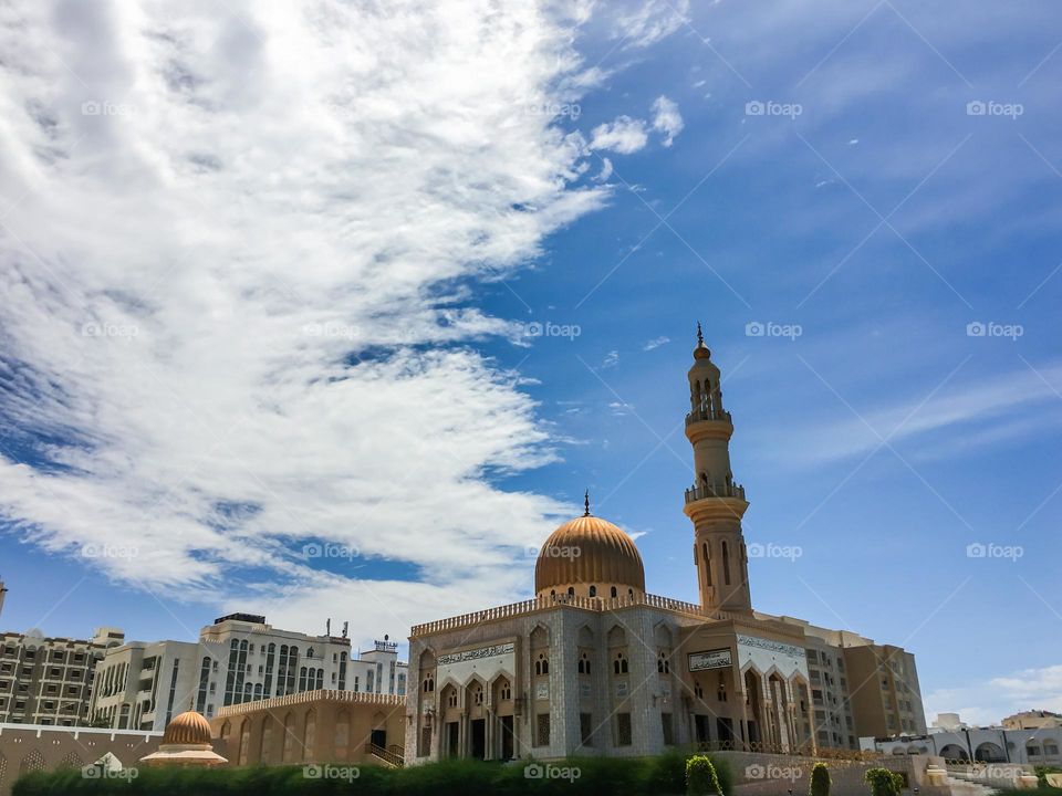 A far shot of a mosque with a central golden dome and minaret, white clouds in the background and a sunny sky