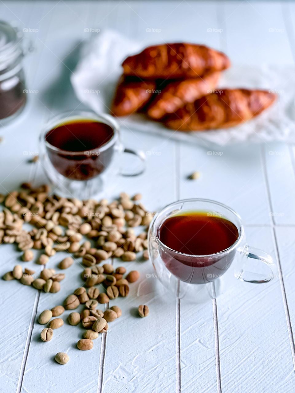 Hot black coffee in a clear cup and coffee beans scattered on top of white table and some croissants at the back
