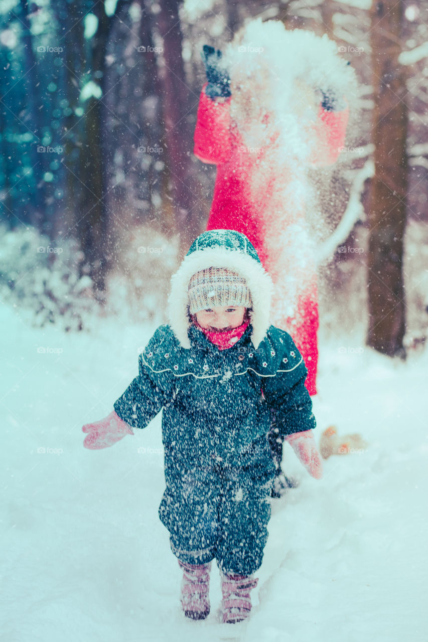 Mother and her little daughter are spending time together walking outdoors in forest in winter while snow falling. Woman is pulling sled, a few years old girl is walking through the deep snow, enjoying wintertime