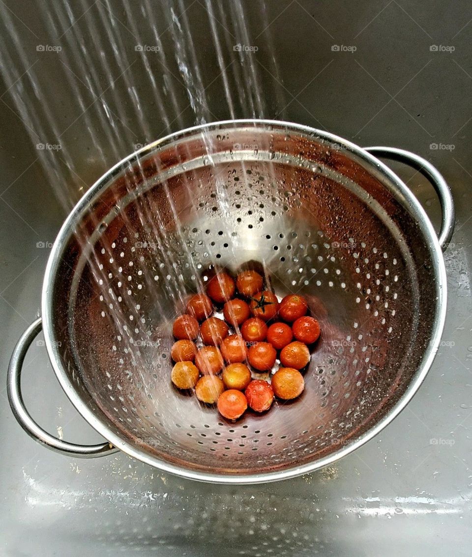 Tomatoes in colander with water spray washing them. They are in sink with water spraying from above.