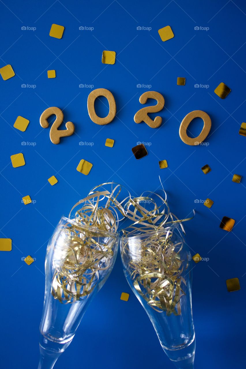 Conceptual flat lay of a champagne toast with glittering gold confetti and the numbers ‘2020’ in gold on a classic blue background - portrait format