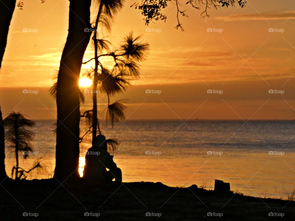 A young lady sits on the bank of the bay absorbing the mesmerizing sunset. The color of twilight and sunset. The dramatic hurried rush of orange collapse as the sun sinks below the gorgeous, cloudy skyline