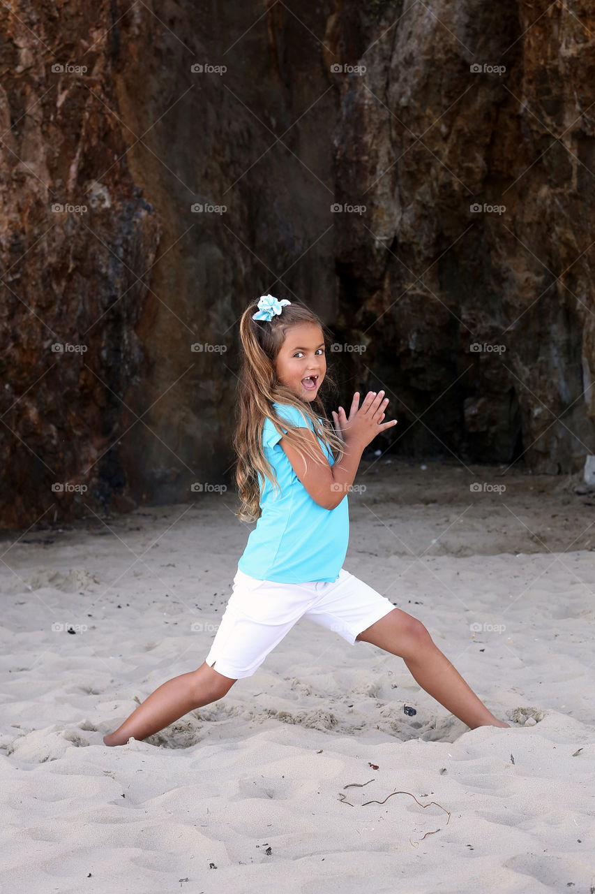 Young girl in teal shirt, white long shorts, and coordinated headband hair tie clasping palms together with giant toothless smile outdoors on beach in front of rock formation. Silly, funny, grin