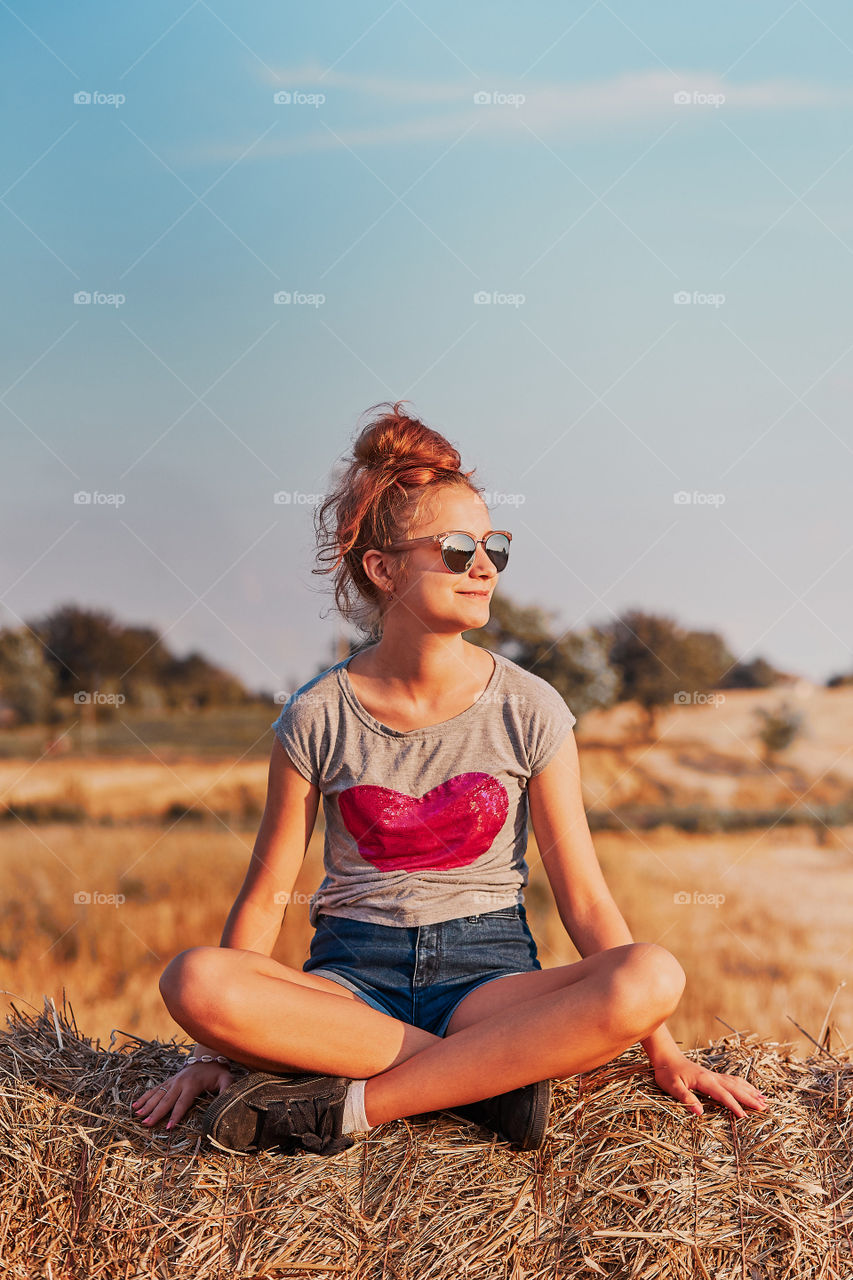 Happy smiling teenage girl sitting on a hay bale at sunset enjoying summer vacations in the countryside. Candid people, real moments, authentic situations