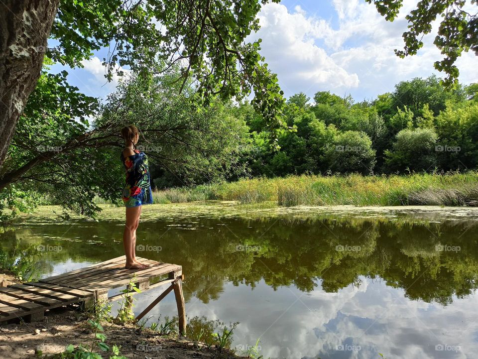 Battle Summer vs. Spring.   The girl stands on a wooden pier on the river bank. Summer on the river
