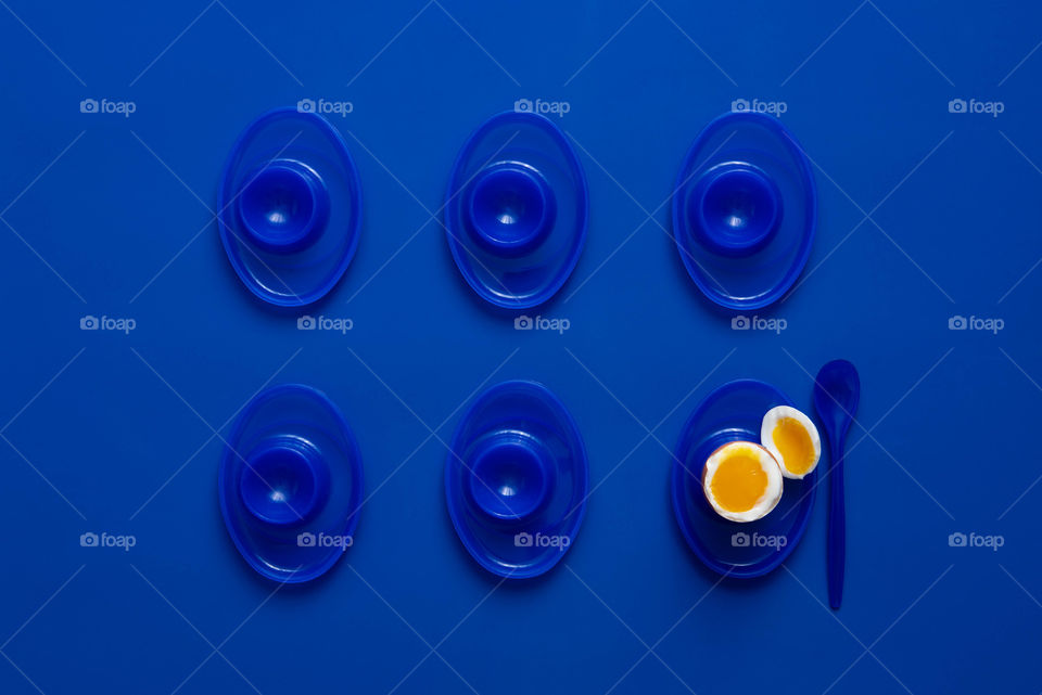 Flat lay of two rows of blue egg cups on a blue background with a single yellow soft boiled egg 