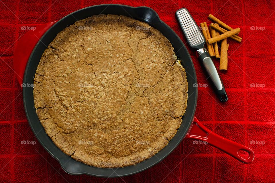 Overhead view of a freshly baked Cinnamon Sourdough Dutch Baby in a red cast iron skillet with cinnamon sticks and mini-grater on a red towel