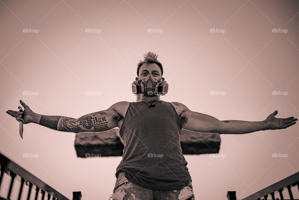Punk man with knife, gas mask and tattoo, standing in front of a cross, black and white