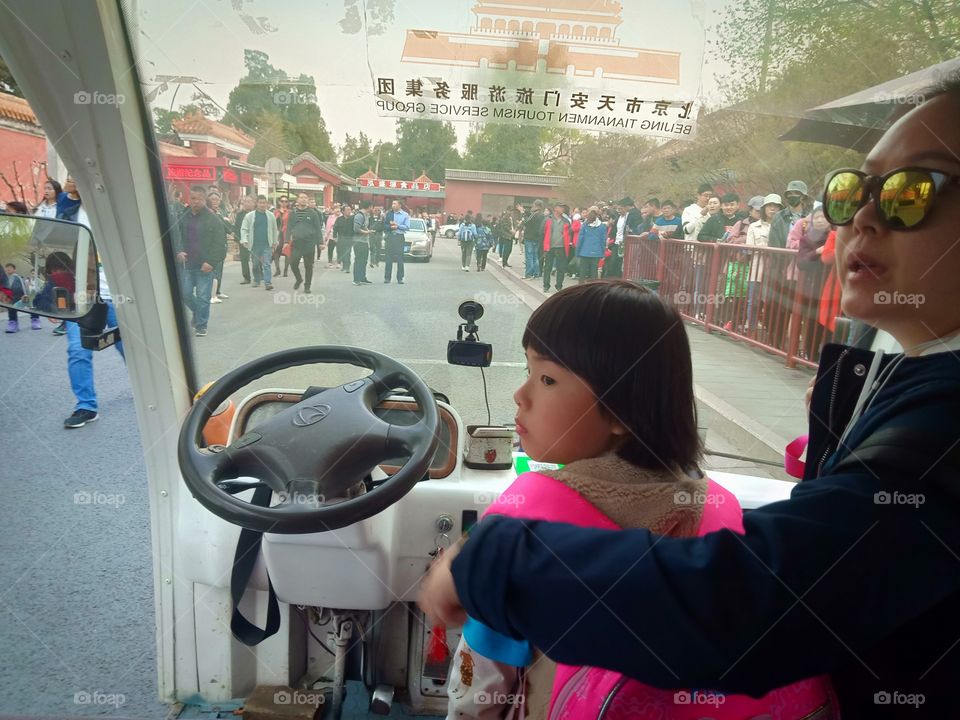 A child with her mother beside turned her head to see outside view while inside passenger carry bus and protective Glass behind.