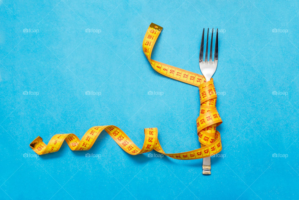 Tape measure around a fork as concept for diet. Fork are wrapped in yellow measuring tape on blue background