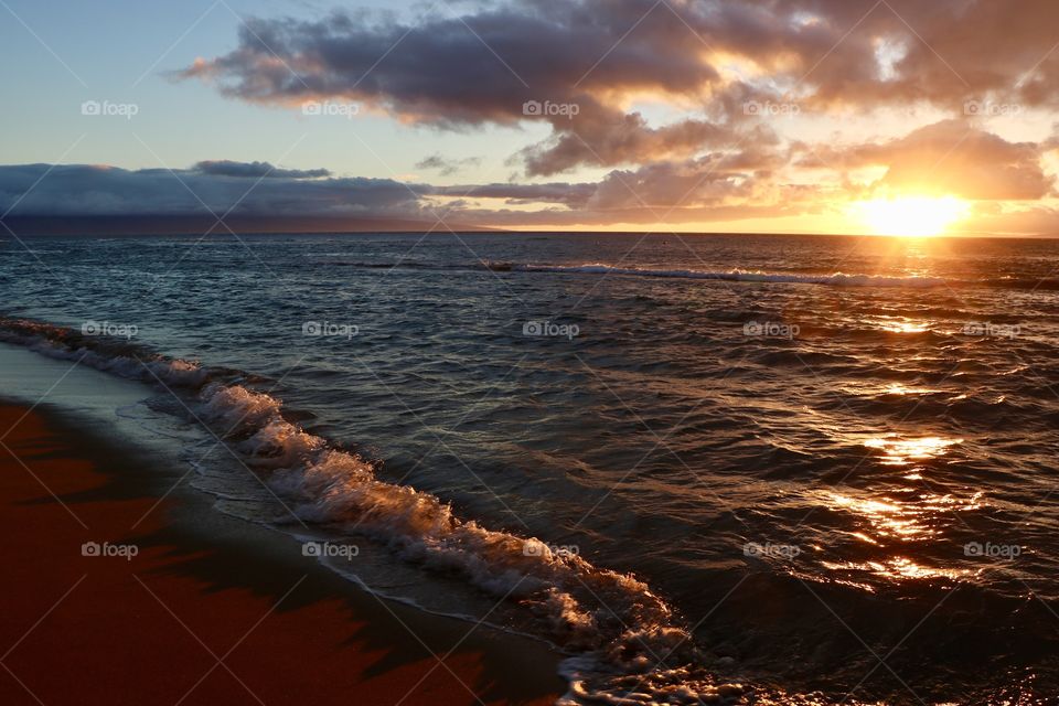 Sun is setting slowly under the clouds while waves are crashing on a sandy beach 