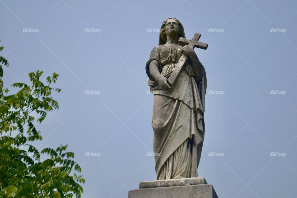 A grey concrete statue of a woman with multi-layered clothing and a head scarf holding a cross on a pedestal against a blue sky